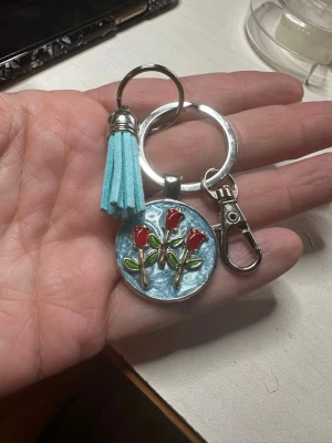 Nyckelring/väsksmycke med röda rosor och tofs - Säljer en rund nyckelring /väsksmycke med blå bakgrund och tre röda rosor i emalj. Nyckelringen har en ljusblå tofs i mockaimitation och en metallisk karbinhake. Perfekt att hänga på väskan eller nycklarna för en personlig touch.