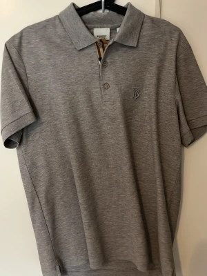 Grå pikétröja från Burberry - Säljer en stilren grå pikétröja från Burberry, perfekt för sommaren och använt max 3 gånger.