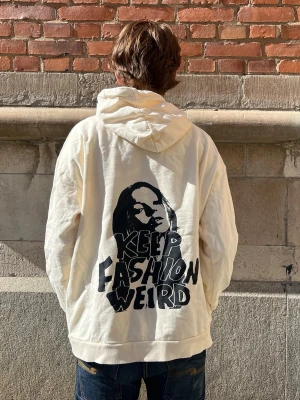 Vit hoodie med tryck och huva -  En oversized vit hoodie med stor huva och känguruficka framtill. På ryggen finns ett stort svart tryck med ett ansikte och texten 'KEEP FASHION WEIRD' i grafisk stil. Hoodien  är gjord i bomullstyg. Perfekt för dig som gillar trender men vill sticka ut lite extra🫰🏻Någr fläckar finns på sidan av tröjan i svart fråga om bild!❤️