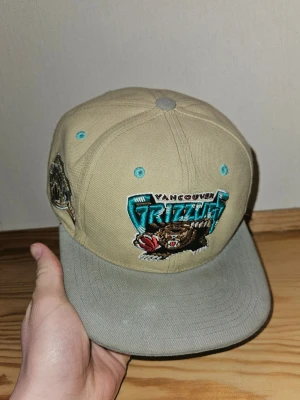 Vancouver Grizzlies beige keps - Sällsynt vintage Vancouver Grizzlies snapback från Mitchell & Ness. Klassiskt 90-tal/early 2000-tal NBA-keps med stort broderat Grizzlies-logo på framsidan och extra patch på sidan. Beige/grå med turkosa detaljer – en riktig grail för NBA-fans och samlare. Justerbar storlek med snapback. Skick: Bra skick inom Vinted – väl använd men fortfarande snygg. Lite naturligt slitage från användning, inga stora skador eller fläckar. Den här är vintage och svår att få tag i längre. Packas säkert och noga. 