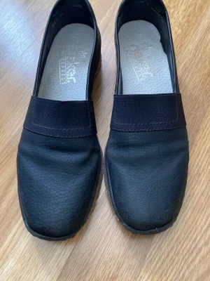 Svarta loafers från Rieker, storlek 38 - Säljer ett par svarta loafers från Rieker i storlek 38. Skorna har en stilren design med elastiskt band över vristen, ovandel i slätt skinn och en bekväm innersula antistress används en gång 