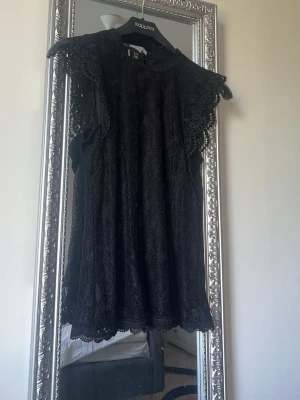 Svart spetsblus från Kappahl - Säljer en svart blus från Pieces med vackra spetsdetaljer och korta ärmar. Blusen har en feminin och luftig design med fina volangkanter vid ärmar och nederkant. Perfekt att styla med jeans eller kjol för en trendig look.