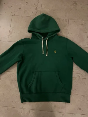 Grön hoodie från Polo Ralph Lauren - Snygg grön hoodie från Polo Ralph Lauren med klassisk gul logga på bröstet. Hoodien har vit dragsko i huvan och en stor magficka framtill. Tillverkad i mjukt material och har ribbade muddar vid ärmslut och nederkant. Perfekt för en avslappnad stil. Hoodien är i otroligt bra skick utan hål eller andra defekter. Tröjan är i storlek S. Obs färgen kan se lite annorlunda ut beroende på ljus. Om du har frågor eller funderingar är det bara att säga till Mvh Olof.