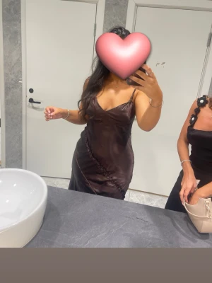 Brun satinklänning med smala axelband - Snygg brun satinklänning med smala axelband och v-ringning. Klänningen har en figurnära passform och en dekorativ rynkdetalj framtill. Längden är midilång och materialet har en glansig finish som ger en lyxig känsla.