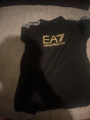 Svart EA7 Emporio Armani t-shirt - Snygg svart t-shirt från EA7 Emporio Armani med guldigt tryck på bröstet och logoband längs axlarna. Klassisk rund hals och korta ärmar. Perfekt för dig som vill ha en stilren och trendig look.