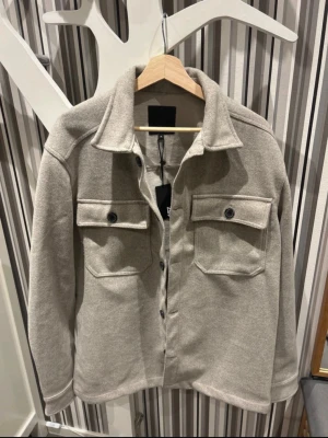 Beige overshirt med fickor, M - Säljer en stilren beige overshirt i mjukt material. Jackan har två stora bröstfickor med knappar, klassisk krage och knäppning framtill. Perfekt lager-på-lager-plagg för dig som gillar enkel och clean stil. Säljer då den ej passar mig. Hör av er för mer frågor😊