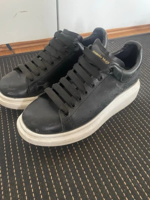 Alexander McQueen svarta sneakers - Svarta sneakers från Alexander McQueen med chunky vit sula och svarta skosnören. Skorna är i läder och har guldtext med logga på plös och häl. Klassisk låg modell med stilren design och perforerade detaljer på sidorna.