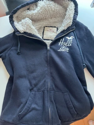 Marinblå Hollister hoodie med foder 2000s zip up - Mysig marinblå hoodie från Hollister med dragkedja och vit broderad logga på bröstet. Insidan är fodrad med fluffigt, vitt teddyfoder och huvan har dragsko. Två fickor framtill gör den både snygg och praktisk. Perfekt för kalla dagar.