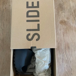 Adidas Yeezy Slide Onyx - Säljer ett par Adidas Yeezy Slide i färgen Onyx. Slidesen har en chunky, vågig sula och är helt svarta. Tillverkade i syntetmaterial och perfekta för en avslappnad stil. Kommer i originalkartong. OBS! Små storlek, passar 39/40. Nypris: 2299kr. 
