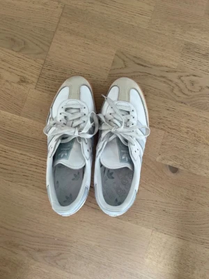 Adidas Samba vita sneakers - Klassiska Adidas Samba sneakers i vitt läder med gråa detaljer och de ikoniska tre ränderna på sidan. Skorna har en låg profil och gummisula som ger en retrovibe. Perfekta för dig som gillar stilrena och tidlösa sneakers.