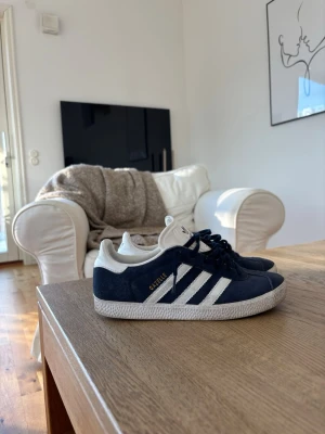 Adidas Gazelle mörkblå sneakers - Säljer ett par klassiska Adidas Gazelle sneakers i mörkblå mocka med vita detaljer och de ikoniska tre ränderna på sidan. Skorna har vit sula och Adidas-logga på hälen. Perfekta för dig som gillar stilrena och tidlösa sneakers.