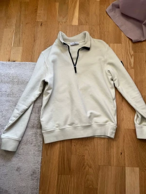 Beige half zip från Stone Island - Snygg beige sweatshirt från Stone Island Junior med svart half zip-dragkedja och hög krage. Tröjan har ribbade muddar vid ärmslut och nederkant samt en klassisk logga på ärmen. Perfekt för en avslappnad och stilren look. Extra knapp vid QR kod. Storlek 12 (XS) men passar större.