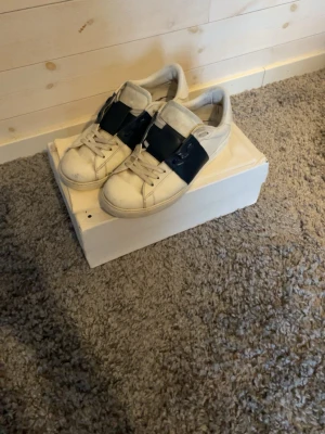 Vita sneakers  - Snygga vita sneakers med ett brett svart band över vristen istället för klassiska snören. Skorna har en stilren och modern design med rund tå och låg profil. Perfekta för dig som gillar en clean look med en twist.