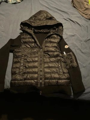 Svart pufferjacka från Moncler 160cm -170cm - Svart pufferjacka från Moncler med huva, ribbstickade ärmar och detaljerad ficka med dragkedja på ena armen. Jackan har quiltad front, två fickor med knappar och coolt Moncler-märke på ärmen. Perfekt för dig som vill ha en snygg och varm jacka med street-vibe. Går att ta telefon mot moncler märket också kommer det upp monclers hemsida passar 160-170cm