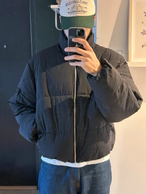 Svart pufferjacka med hög krage - Säljer en svart pufferjacka med hög krage och dragkedja framtill. Jackan har en boxig passform och är perfekt för kalla dagar. Fodrad och riktigt varm, passar dig som gillar en stilren och enkel look. Den är aldrig använd och kommer med prislappen fortfarande på och inkluderar en avtagbar luva. Ordinarie pris 899kr