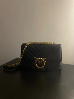 Svart handväska - Snygg svart quiltad handväska med guldfärgad kedjerem och ikoniskt fågelspänne framtill. Väskan har en stilren rektangulär form och elegant mönster i material som ser ut som läder. Perfekt accessoar för att lyfta din outfit.