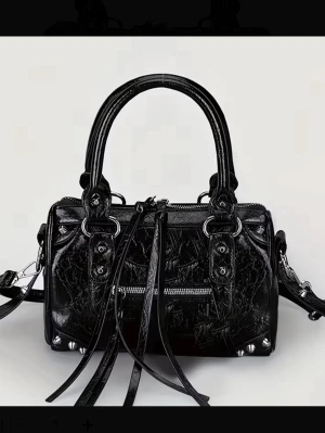 city bag - Cool svart handväska i glansigt konstläder med silverfärgade metalldetaljer, nitar och dragkedjor. Väskan har dubbla handtag, avtagbar axelrem och långa dekorativa remmar framtill. Perfekt för dig som gillar edgy och trendig stil.