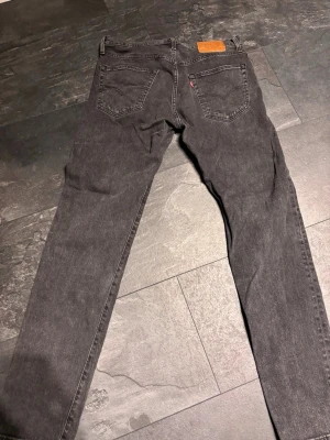 Grå Levi's jeans raka ben - Säljer ett par grå Levi's jeans med raka ben och klassisk femficksdesign. Jeansen har en liten röd Levi's-tag på bakfickan och läderpatch i midjan. Tillverkade i slitstark denim, perfekta för en avslappnad och stilren look.