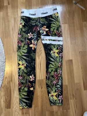 Stronger leggings med blommigt mönster - Snygga leggings från Stronger med tropiskt blommigt mönster i grönt, gult, rosa och vitt. Bred vit resår i midjan med logga och två vita ränder på ena benet. Perfekta för träning eller chill. Stretchigt material som sitter skönt.