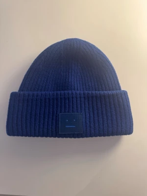 Acne Studios mössa - Säljer en blå ribbstickad mössa från Acne Studios i 100% ull. Mössan har uppvikt kant och Acne Studios klassiska ansiktslogga framtill i matchande blått. Mössan har tvättats och därmed krympts därav sitter loggan lägre ner än vad den ska och därav priset. Priset är diskuterbart vid snabb affär!