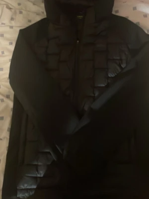 Svart pufferjacka från Everest - Säljer en svart pufferjacka från Everest med quiltad design och huva. Jackan har dragkedja framtill och diskret logga på bröstet. Perfekt för kalla vinterdagar tack vare det tjocka, vadderade materialet som håller värmen.