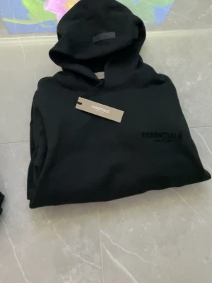 Svart Essentials hoodie med huva - Snygg svart hoodie från Essentials med diskret logga på bröstet och klassisk huva. Hoodien har en minimalistisk design och är tillverkad i mjukt material som känns skönt mot huden. Perfekt för dig som gillar streetwear och enkel stil.