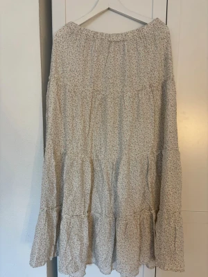 Lång blommig volangkjol i bomull - Söt långkjol med volanger i ljusbeige bomullstyg med småblommigt mönster i rosa och grönt. Kjolen har en avslappnad passform och resår i midjan för extra komfort. Perfekt för dig som gillar romantisk och bohemisk stil.