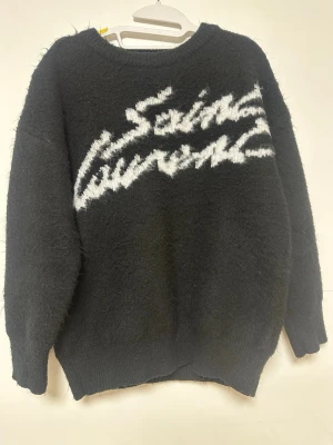 Svart Saint Laurent stickad tröja - Mysig svart stickad tröja från Saint Laurent med vit logotyp i grafisk stil över bröstet. Tröjan har rund halsringning, långa ärmar och ribbade muddar. Materialet ser fluffigt och mjukt ut, perfekt för kalla dagar.