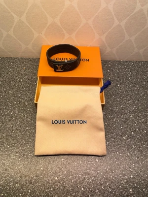 Louis Vuitton svart läderarmband - Snyggt svart armband från Louis Vuitton i präglat läder med det klassiska LV-märket i silver framtill. Armbandet har monogramdetaljer och kommer med original ask och tygpåse. Perfekt accessoar för dig som gillar exklusiv och stilren design. Men ! Den har en liten skada som ni ser på sista bilden.  Men den går att använda om man är försiktig 😁