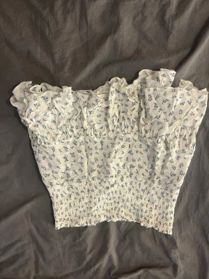 Vit blommig volangkjol - Supersöt kort kjol i vit med små blå blommor och volang upptill. Kjolen har smock i midjan för en bekväm passform och ett romantiskt, somrigt uttryck. Perfekt för dig som gillar en feminin och trendig stil.