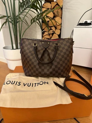Louis Vuitton Siena MM - Snygg handväska från Louis Vuitton i klassiskt brunt Damier-mönster. Väskan har två handtag, avtagbar axelrem och guldfärgade detaljer. Insidan är fodrad i rött tyg med flera fack för enkel organisering. Perfekt för dig som gillar lyxiga accessoarer. I god skick , använt 2-3 gånger ! Dustbag,kartong och kvitto finns . 