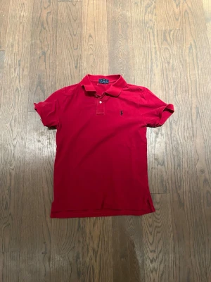 Röd pikétröja från Polo Ralph Lauren - Klassisk röd pikétröja från Polo Ralph Lauren med broderad logga på bröstet. Tröjan har korta ärmar, krage och två knappar vid halsen. Tillverkad i mjuk bomull och har en normal passform. Perfekt för en avslappnad men stilren look. Passar 14 år