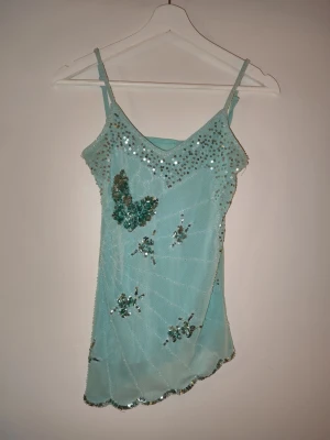 Y2K Ljusblå paljettlinnetopp med fjärilar - Supersöt ljusblå linnetopp med tunna axelband och glittriga paljettdetaljer. Toppen har ett fjärilsmotiv och flera små blommor i paljetter, samt vågig nederkant. Perfekt för dig som vill sticka ut med en unik och trendig topp. Bedömer storleken till cirka XS/ liten S. Färgen är en typ av turkosblå 