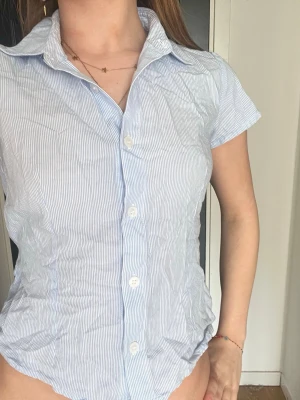 Blårandig skjorta med knyt i midjan - Snygg blårandig skjorta med korta ärmar och klassisk krage. Skjortan har knappar framtill och ett knytband i midjan för en mer figurnära passform. Tillverkad i tunn bomull, perfekt för varma dagar.