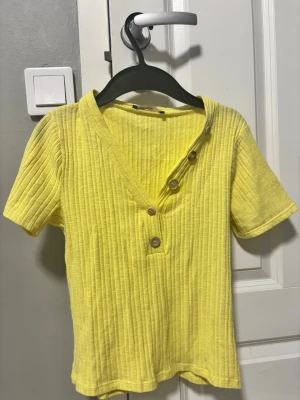 Gul ribbad topp med knappar köpt utomlands  - Snygg gul ribbad topp från H&M med korta ärmar och V-ringning. Toppen har dekorativa träknappar längs halsringningen och är gjord i ett mjukt, stretchigt material som sitter skönt på kroppen. 