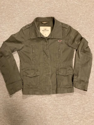 Olivgrön jacka från Hollister - Hollister Jacka/Overshirt i mycket bra skick! Det finns en defekt bak på högerarm vilket är en liten missfärgning kan man säga. se bild 7. För övrigt i väldigt bra skick! Personen på bilden är 180 cm lång, hör av er om frågor!