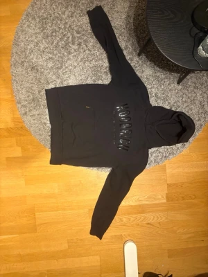 Hoodrich Hoodie Svart - Säljer en svart hoodie från Hoodrich med stor ficka framtill och huva. Hoodien har ett stort, glansigt Hoodrich-tryck över bröstet och är gjord i mjukt bomullsmaterial. Perfekt för en chill och street-inspirerad look.