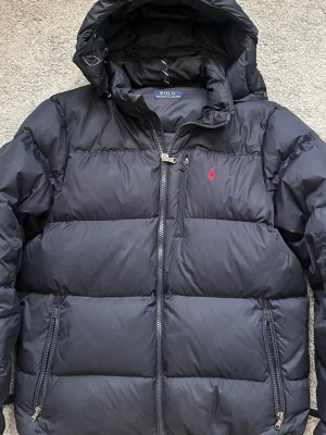 Ralph Lauren Jacka - Säljer en mörkblå Ralph Lauren jacka i fint skick, enbart ett litet hål på ena ärmen (se bild 2). Storlek L: bröstvidd 130cm, axel 53cm, ärm 96,5cm, nederkant 125cm (justerbar).