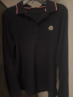 Mörkblå Moncler pikéskjorta - Snygg mörkblå långärmad pikéskjorta från Moncler med klassisk krage och rödvit rand på kragen. Bröstet pryds av Monclers ikoniska logotyp. Perfekt för en stilren och sportig look.