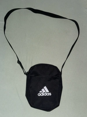 Svart axelväska från Adidas - Snygg svart axelväska från Adidas med justerbar rem och klassisk vit logga framtill. Väskan har dragkedja och ett huvudfack som rymmer det viktigaste. Perfekt för dig som vill ha en sportig och enkel look. Tillverkad i slitstarkt tygmaterial.