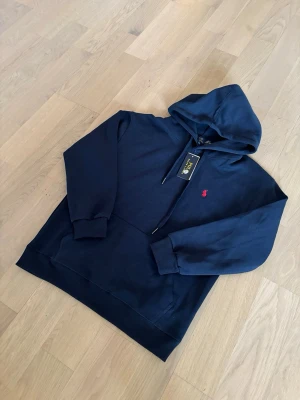 Mörkblå hoodie från Polo Ralph Lauren - Snygg mörkblå hoodie från Polo Ralph Lauren med klassisk huva och dragsko. Tröjan har en liten röd logga broderad på bröstet och ribbade muddar vid ärmar och nederkant. Tillverkad i mjukt bomullsmaterial för skön känsla.