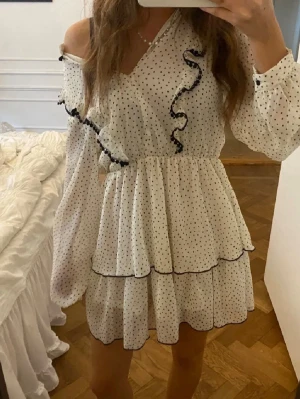 Vit prickig volangklänning med v-ringning - Söt kort volangklänning i vitt med svarta prickar och svarta volangdetaljer längs v-ringningen. Klänningen har långa ärmar och omlottkänsla upptill. Perfekt för dig som gillar romantisk och lekfull stil.
