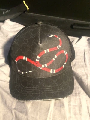 Svart Gucci keps med ormtryck - Snygg svart keps från Gucci med ikoniskt GG-mönster och en röd-vit orm på framsidan. Kepsen har mesh på baksidan för extra ventilation och justerbar passform. Materialet är en mix av bomull och polyester, perfekt för en trendig streetwear-look.