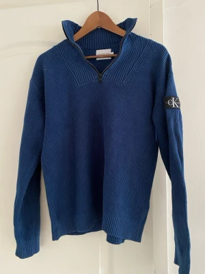 Blå stickad tröja från Calvin Klein - Blå ribbstickad tröja från Calvin Klein med halv dragkedja och hög krage. Tröjan har lång ärm och en svart CK-logga på vänster ärm. Perfekt för dig som gillar stilren och enkel design.