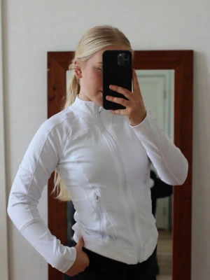 Vit lululemon define jacket - Snygg vit sportjacka från Lululemon med dragkedja framtill och två fickor med dragkedja. Jackan har figurnära passform, hög krage och är tillverkad i ett stretchigt funktionsmaterial som andas. Perfekt för träning eller löpning. Helt ny