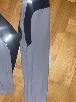 Under armour set - Snyggt träningsset från Under Armour med jacka och byxor i grått och svart. Jackan har hel dragkedja, huva och reflekterande detaljer samt logga på bröstet och ärmen. Byxorna har matchande färgblock och bekväm passform. Perfekt för löpning eller gym.