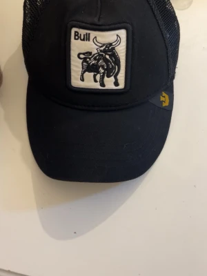 Svart Bull keps med nät och patch - Svart keps från Goorin Bros med vit patch framtill med en tjur och texten 'Bull'. Kepsen har böjd skärm, justerbar snapback och sidor i mesh för extra ventilation. Perfekt för en cool och avslappnad stil.