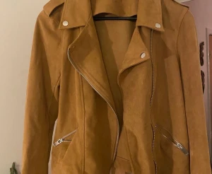 Brun skinnjacka från KappAhl - Snygg brun skinnjacka från KappAhl i bikerstil med silvriga dragkedjor och tryckknappar. Jackan har bred krage och två fickor med dragkedja framtill. Perfekt för dig som vill ha en trendig och stilren look.