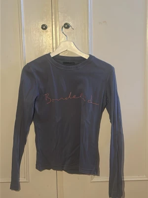 Blå långärmad topp från Bondelid - Mörkblå långärmad topp från Bondelid med rund halsringning och rosa broderad logga framtill. Tillverkad i mjukt bomullsmaterial som sitter skönt mot kroppen. Perfekt till jeans eller kjol för en enkel och snygg look.