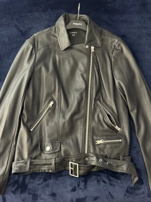 Svart skinnjacka från Lindex - Svart skinnjacka från Lindex med asymmetrisk dragkedja, flera fickor med dragkedjor och bälte i midjan. Klassisk bikerstil med bred krage och silverfärgade detaljer. Perfekt för dig som vill ha en edgy look.